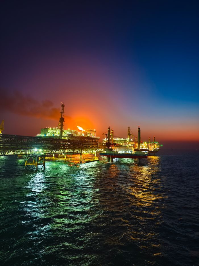 Illuminated offshore oil rig in the ocean during a vibrant sunset, reflecting on the water.